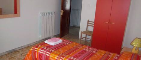 A bedroom with a bed, a red wardrobe, a chair, a radiator, and a mirror on the wall.
