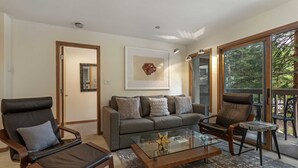 Ein modernes Wohnzimmer mit einer Couch, zwei Ledersesseln, einem Glastisch und einem großen Fenster mit Blick auf Bäume.
