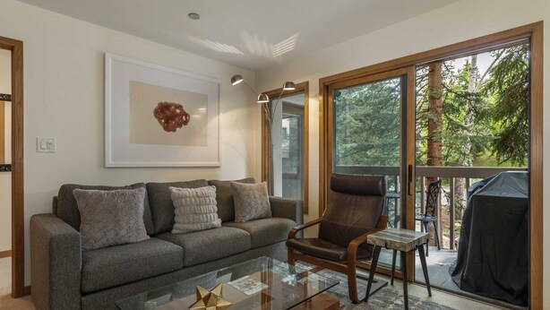 Ein modernes Wohnzimmer mit einem grauen Sofa, einem Holz-Sessel und einem Glastisch. An der Wand hängt ein gerahmtes Kunstwerk und ein großes Fenster bietet Blick auf Bäume.