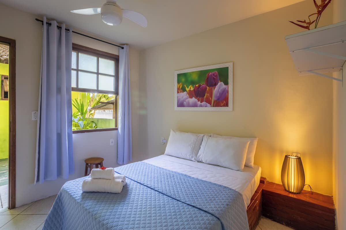 Une chambre d’hôtel avec un lit, une fenêtre avec des rideaux, un petit tabouret, une étagère fixée au mur et un tableau.