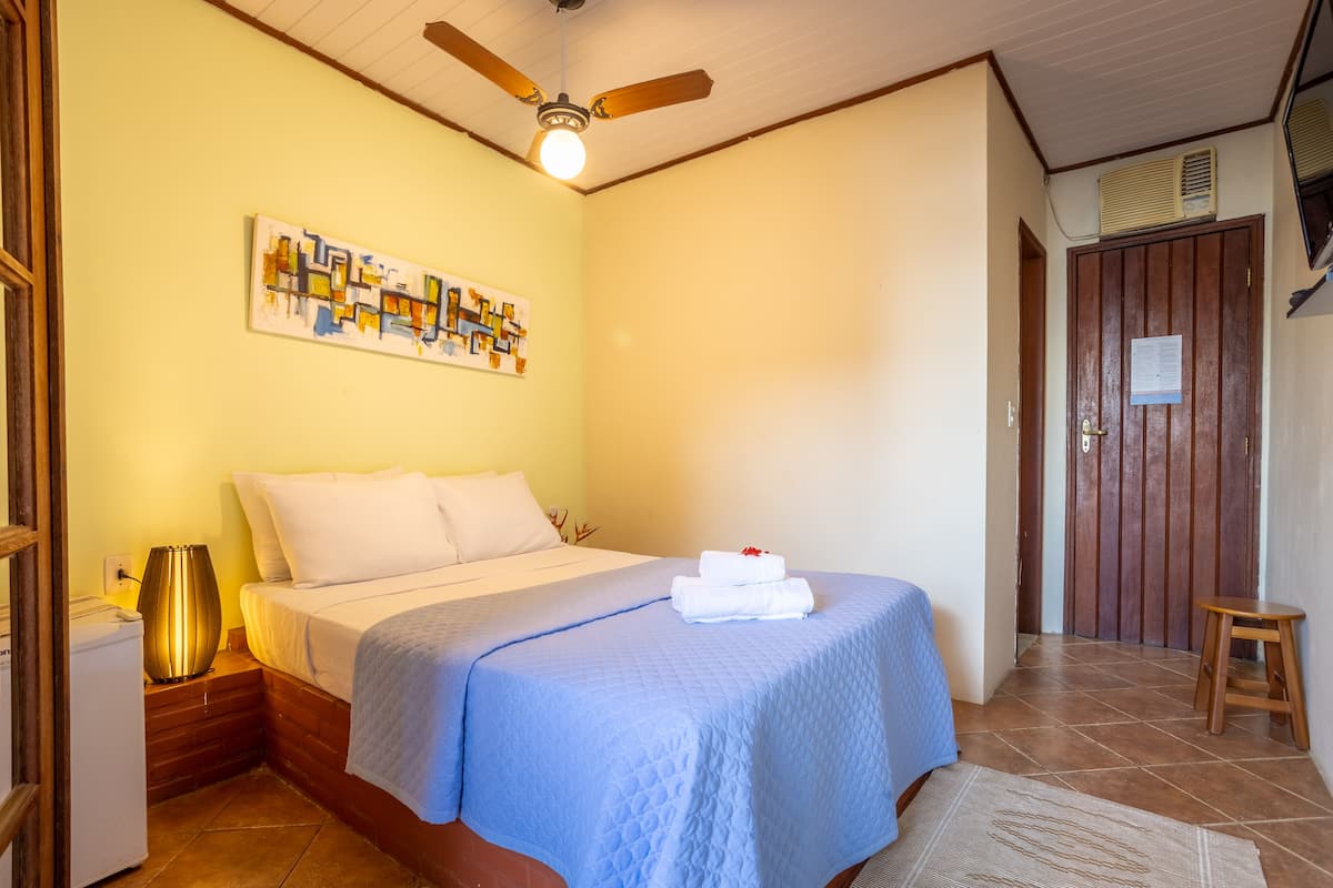 Une chambre d’hôtel avec un lit, un ventilateur de plafond, une œuvre d’art accrochée au mur, une porte en bois et un petit tabouret.