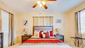 Quarto de hotel com uma cama grande, um ventilador de teto, mesa de cabeceira com abajur e janela com cortinas.