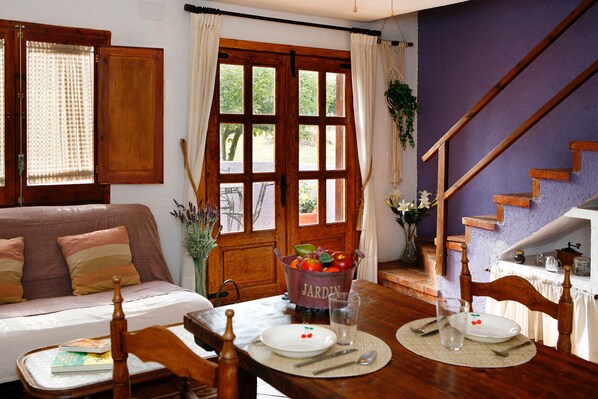 A dining area with a wooden table set for two, a wooden chair, a bowl of fruit, and a view of a staircase. - Self catering La Ginesta rural for 20 people (Sitges)