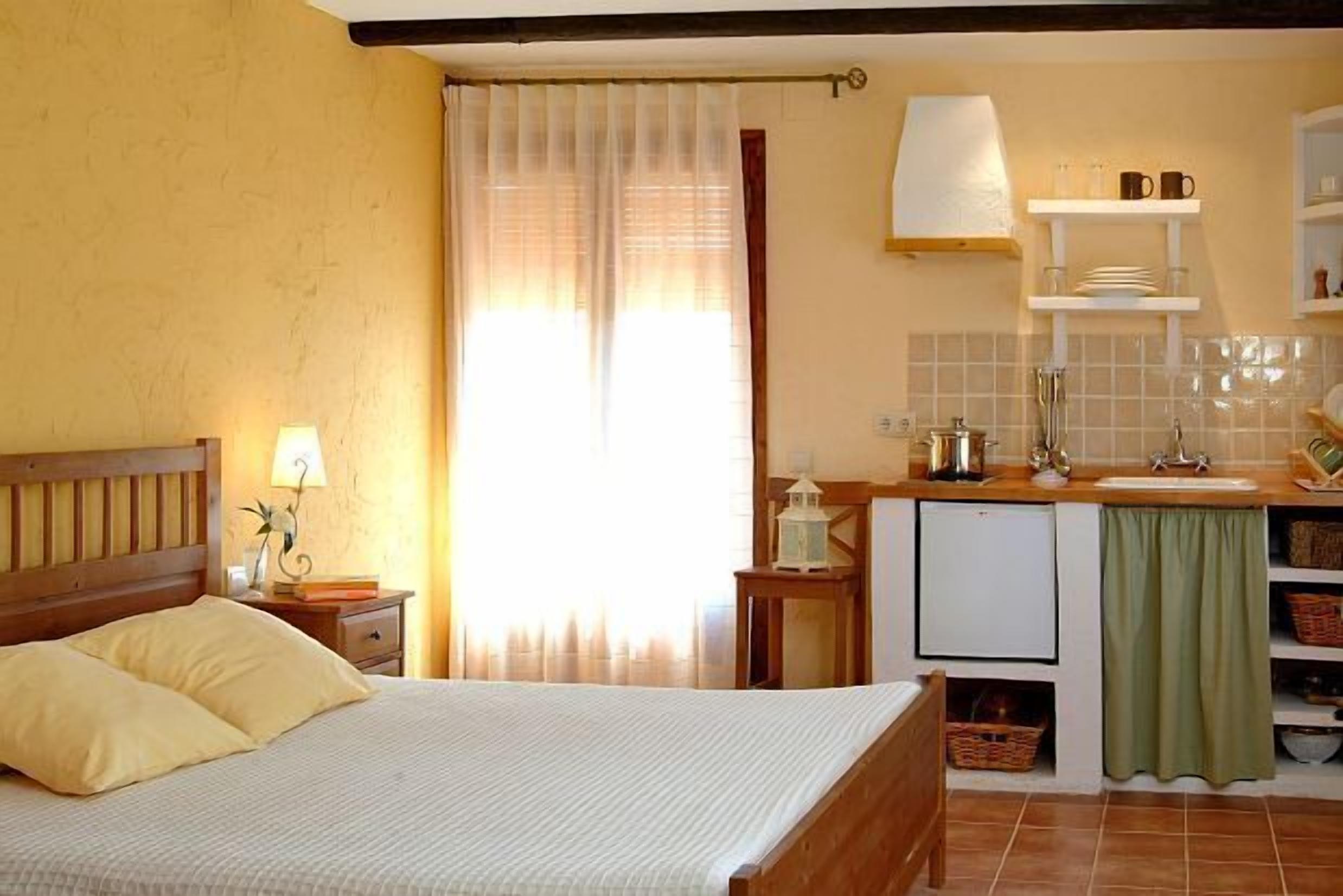Een slaapkamer met een bed, nachtkastje, lamp, en uitzicht op een keuken met een spoelbak, kastjes en een raam met gordijnen.