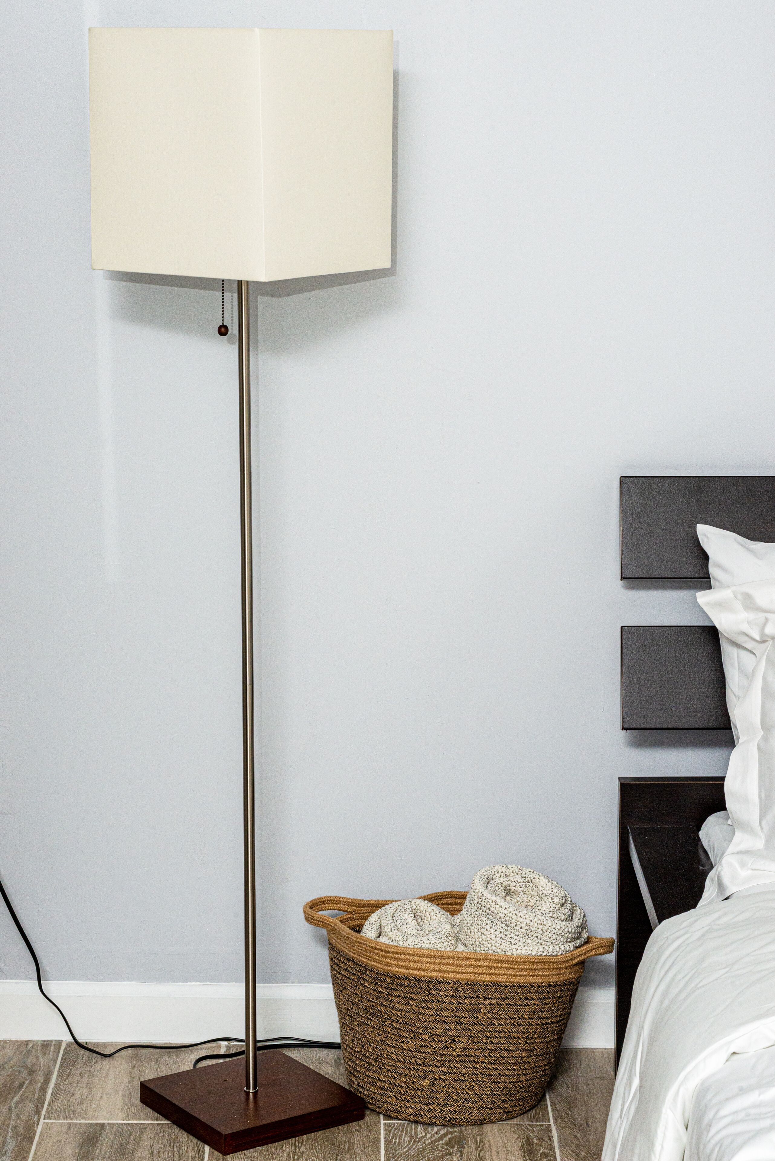 Eine Stehlampe mit weißem Lampenschirm, ein gewebter Korb mit Decken und ein Bett mit weißer Bettwäsche.