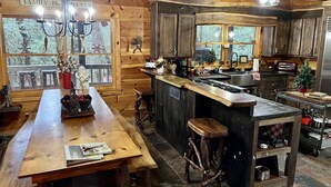A cozy cabin-style kitchen with wooden walls, a central island with stools, a chandelier, and a view of the forest through large windows.