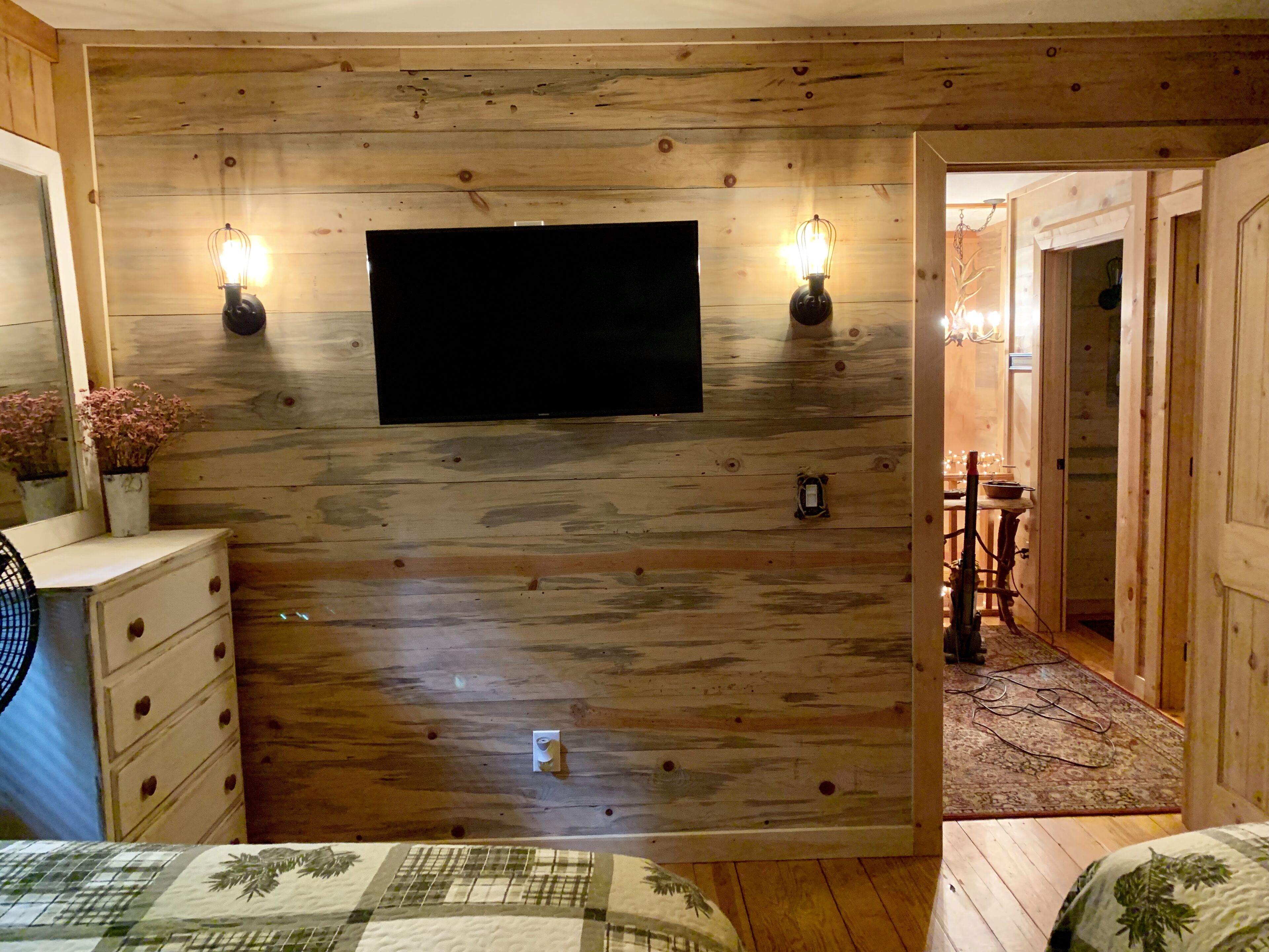 A wooden-paneled room with a flat-screen TV mounted on the wall, two wall sconces, a dresser, a bed with a patterned bedspread, and a hallway leading to another room.