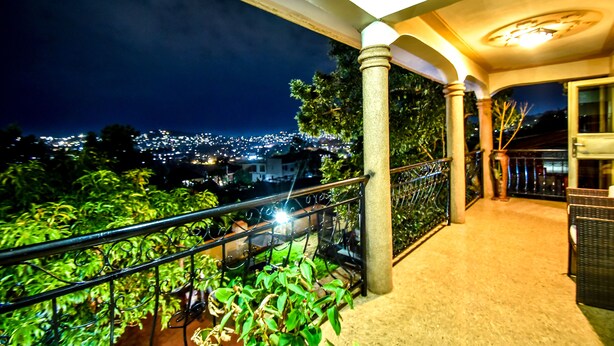Un balcon avec une balustrade, une vue sur le paysage urbain de nuit, et un bâtiment doté de colonnes et d’une porte.
