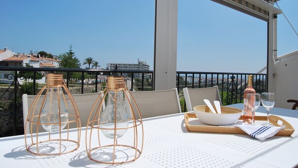 Une table dressée pour deux personnes, avec une bouteille de vin rosé, deux verres à vin, un bol et deux chandeliers, sur un balcon donnant sur un paysage urbain.