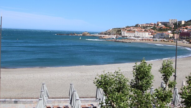 A coastal view with a beach, a rocky breakwater, and buildings on a hillside.
