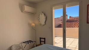 A bedroom with a bed, a chair, a lamp, an air conditioner, and a sliding glass door leading to a balcony with a tiled floor and a view of buildings.