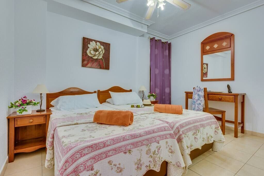 A bedroom with a double bed, two bedside tables, a nightstand, a desk, a chair, a mirror, and a ceiling fan.