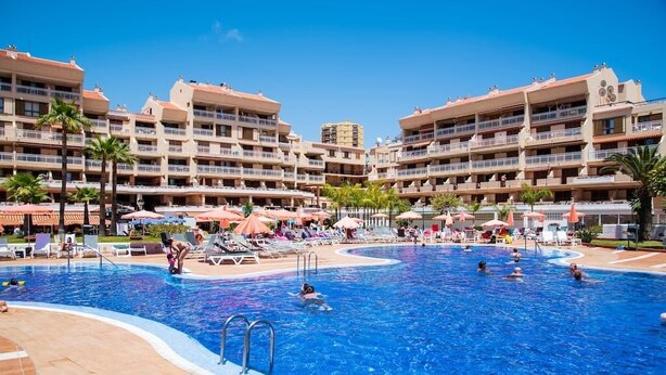 Uma grande piscina cercada por edifícios de hotel com várias camadas, varandas, espreguiçadeiras e guarda-sóis.