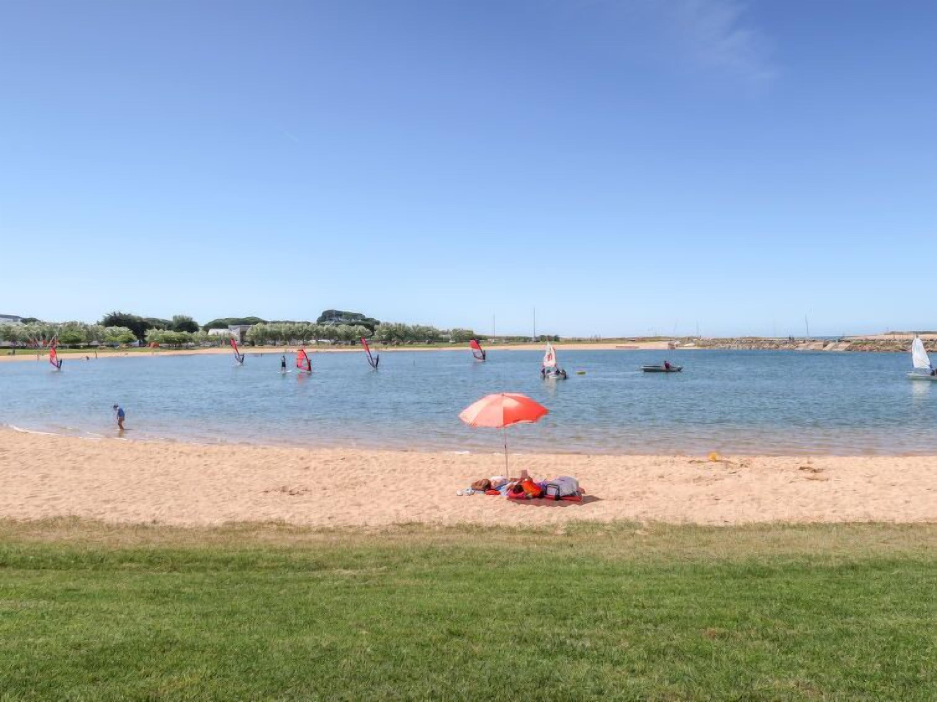 Une plage où l’on voit des gens pratiquer des activités nautiques, un parasol rouge et des bateaux sur l’eau.