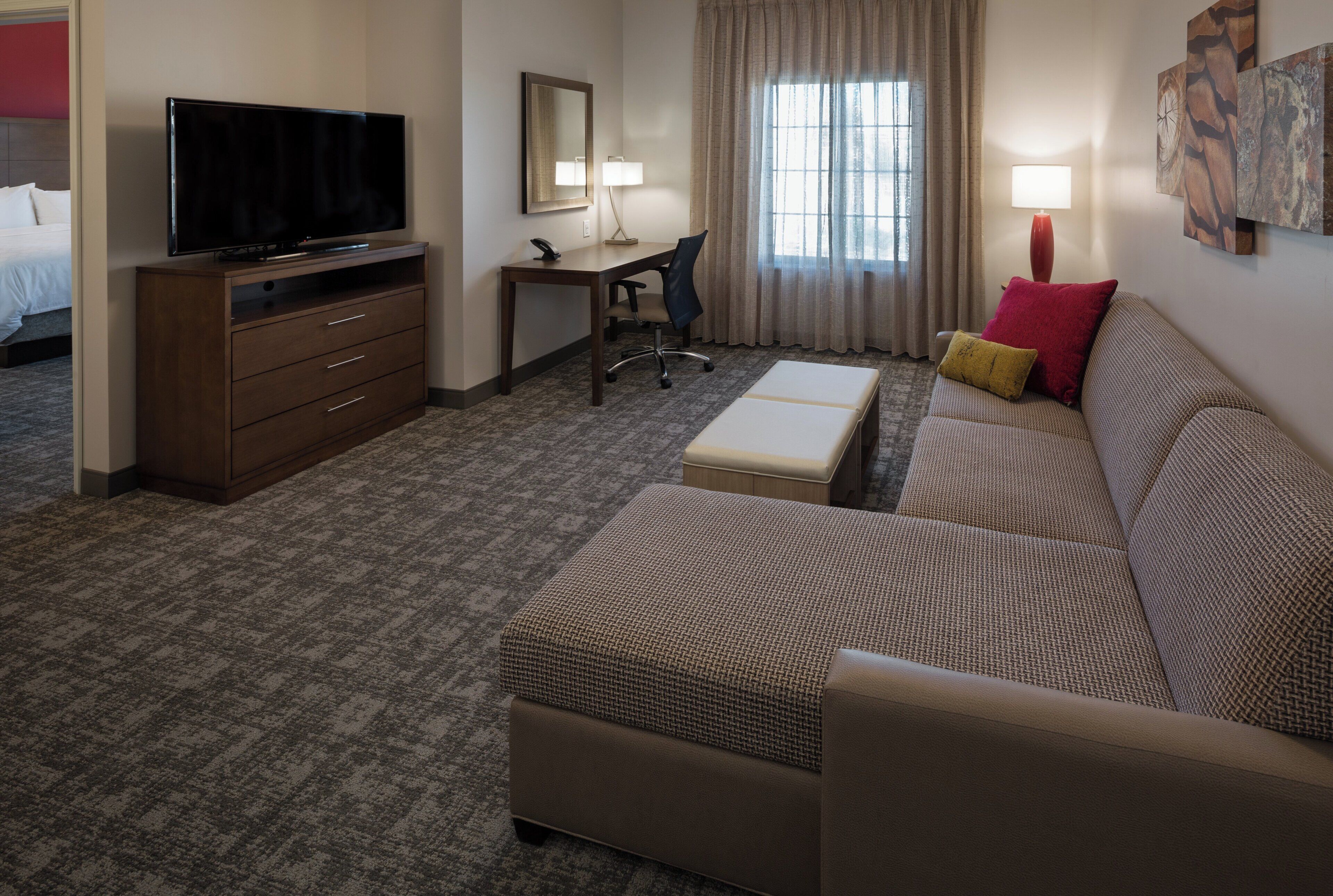 A hotel room with a flat-screen TV, a desk with a chair, a sofa with a footrest, and a window with curtains.