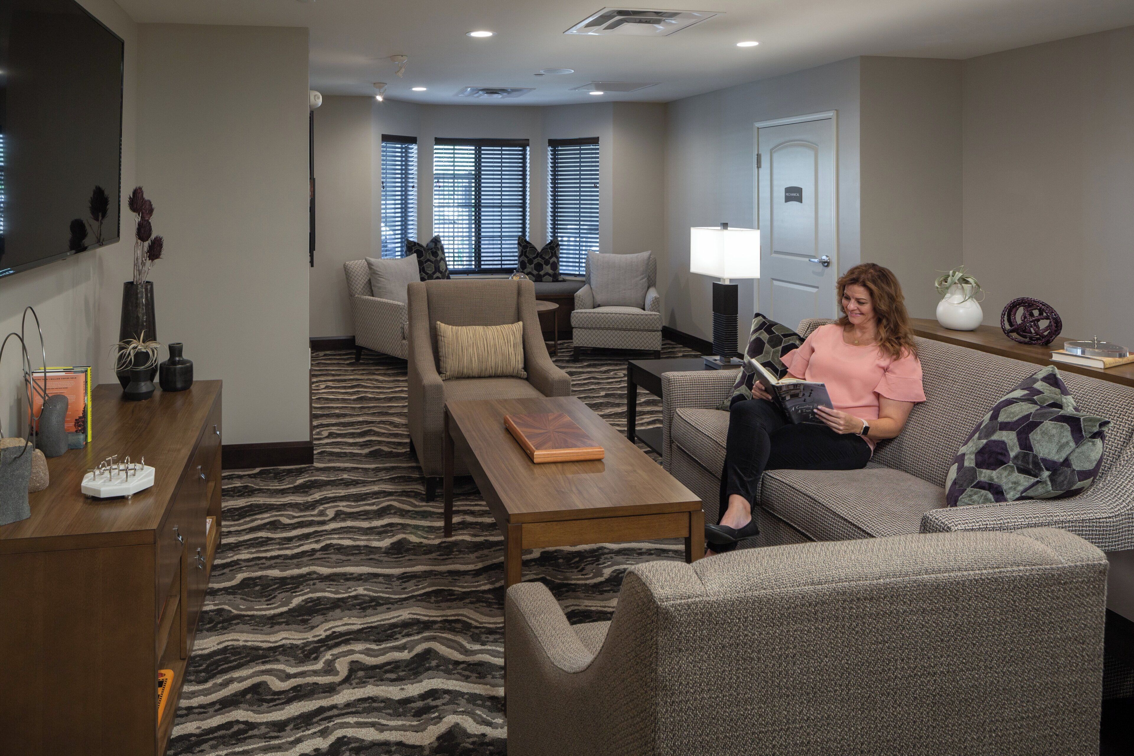 A modern living room with a sofa, armchair, and coffee table. There is a television mounted on the wall, a door labeled 'Technical,' and a window with blinds.