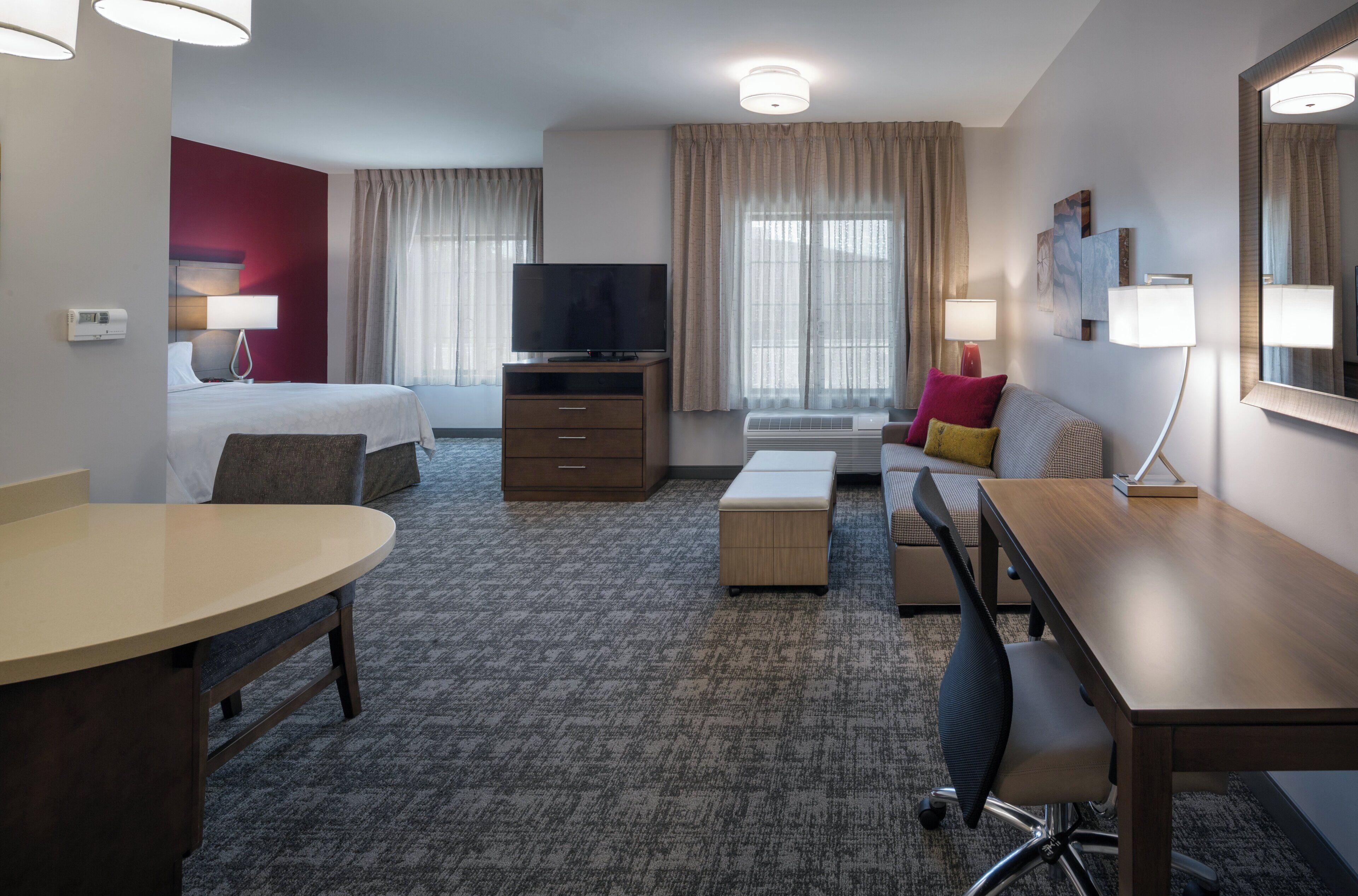 A hotel room with a bed, a desk, a chair, a TV, a lamp, and a window with curtains.