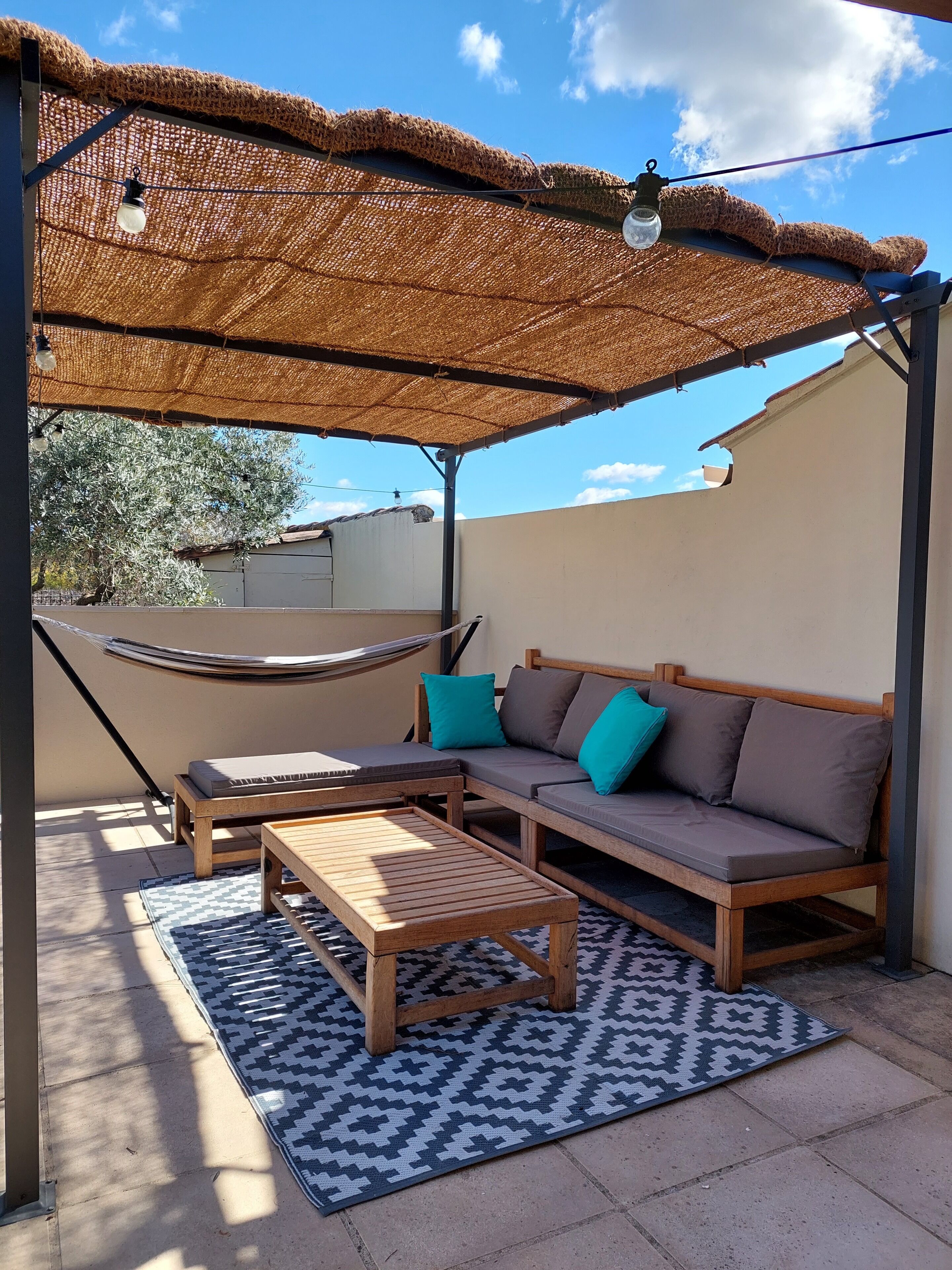 An outdoor seating area with a thatched roof, a wooden sofa with cushions, and a patterned rug.