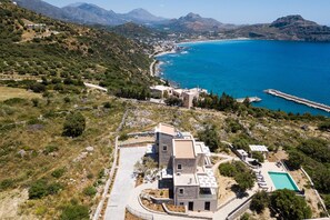 An aerial view of a coastal property with a swimming pool, a paved driveway, and a view of the sea and mountains.
