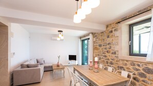 A modern living room with a sofa, a dining table, and chairs. There is a TV on a stand and a stone wall. - Endless Blue View, Top Beaches Nearby (Rethymno)
