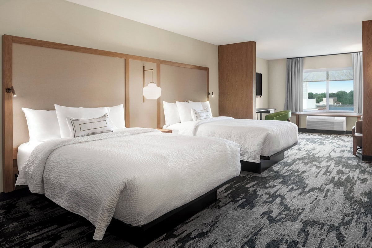 A hotel room with two beds, a large window with curtains, a wall-mounted TV, and a carpeted floor.