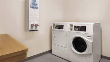 A laundry facility with a wall-mounted soap and bleach dispenser, two stacked front-loading washing machines, and a wooden bench.