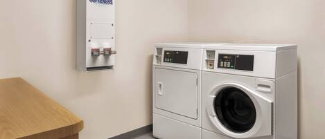 A laundry facility with a wall-mounted soap and bleach dispenser, two stacked front-loading washing machines, and a wooden bench.