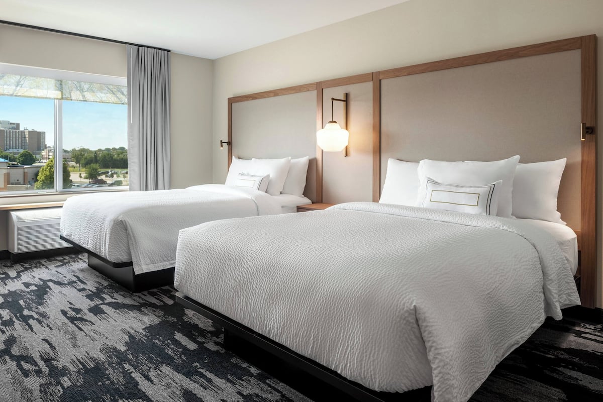 A hotel room with two beds, a large window with a view of buildings, and a patterned carpet.