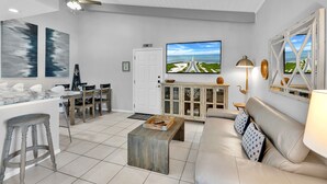 A modern living room with a large TV, a wooden coffee table, a sofa, and a dining area with chairs and a cabinet. - New Listing! Beachside gem, newly remodeled & redecorated  (South Padre Island)