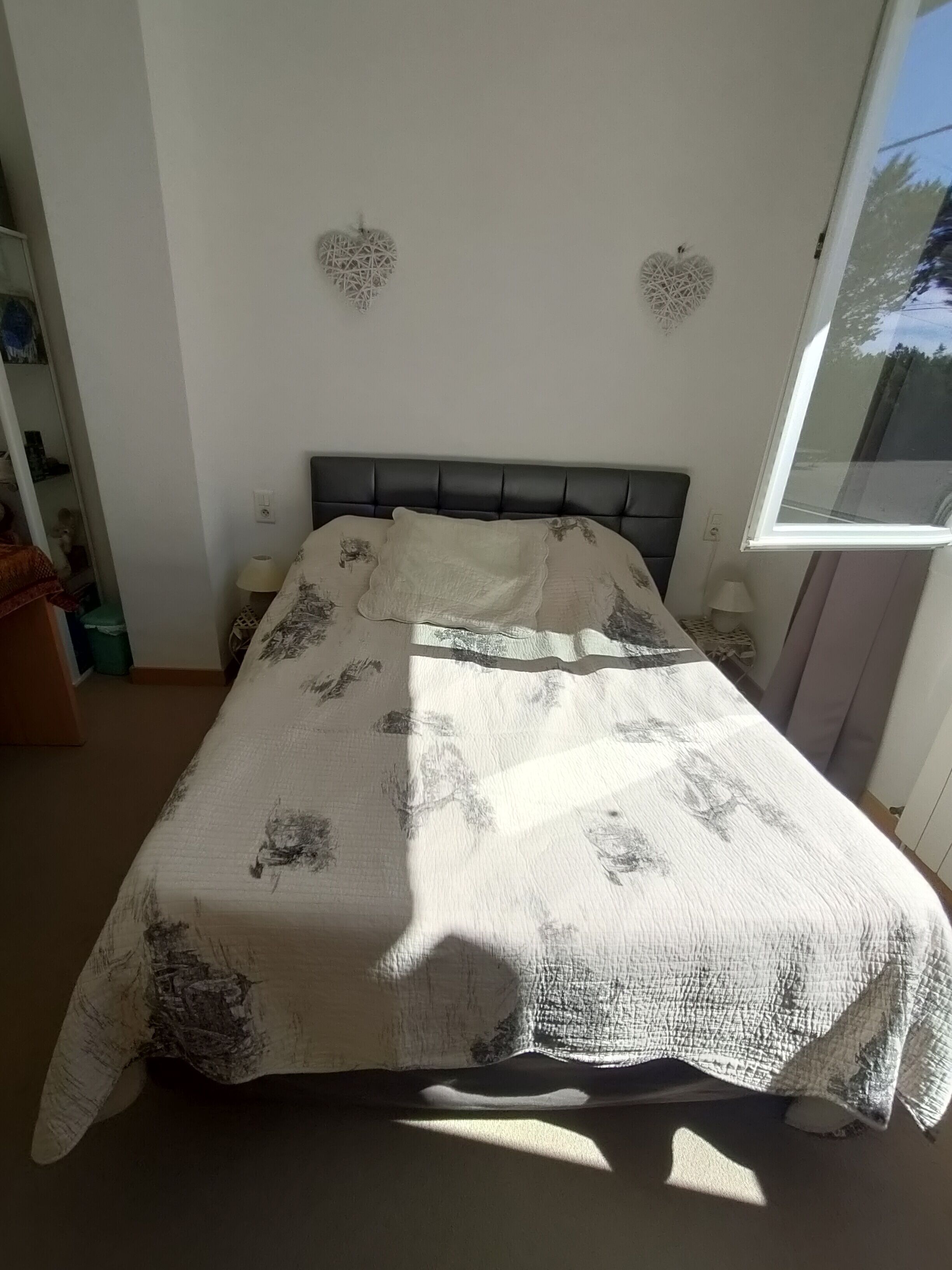 Un lit recouvert d’une couvre-lit à motifs, deux décorations en forme de cœur fixées au mur, une fenêtre et une table de chevet avec une lampe.