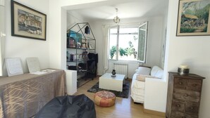 A living room with a sofa, a coffee table, a bean bag, a bookshelf, and a window with a view of greenery.