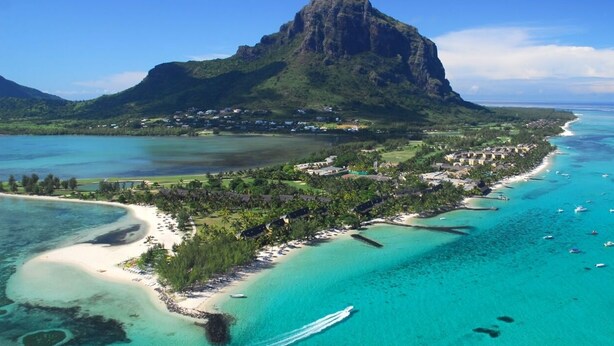 Vue aérienne d’une station balnéaire, avec une montagne imposante en arrière-plan, des eaux turquoise limpides et une plage où se trouvent plusieurs bateaux.