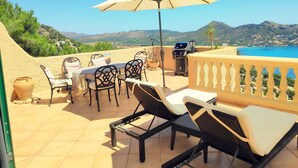 Un patio al aire libre con una mesa, sillas y una parrilla, con vistas a un paisaje pintoresco que incluye montañas y una masa de agua.