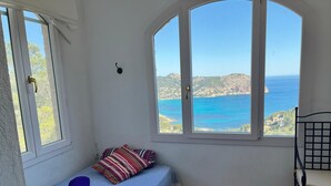 A small room with a bed, a chair, and a large window offering a view of the sea and hills.