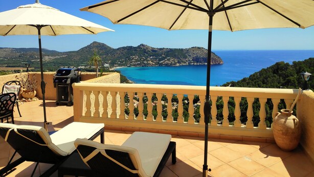 A terrace with two sun umbrellas, a grill, and a view of the sea and mountains.