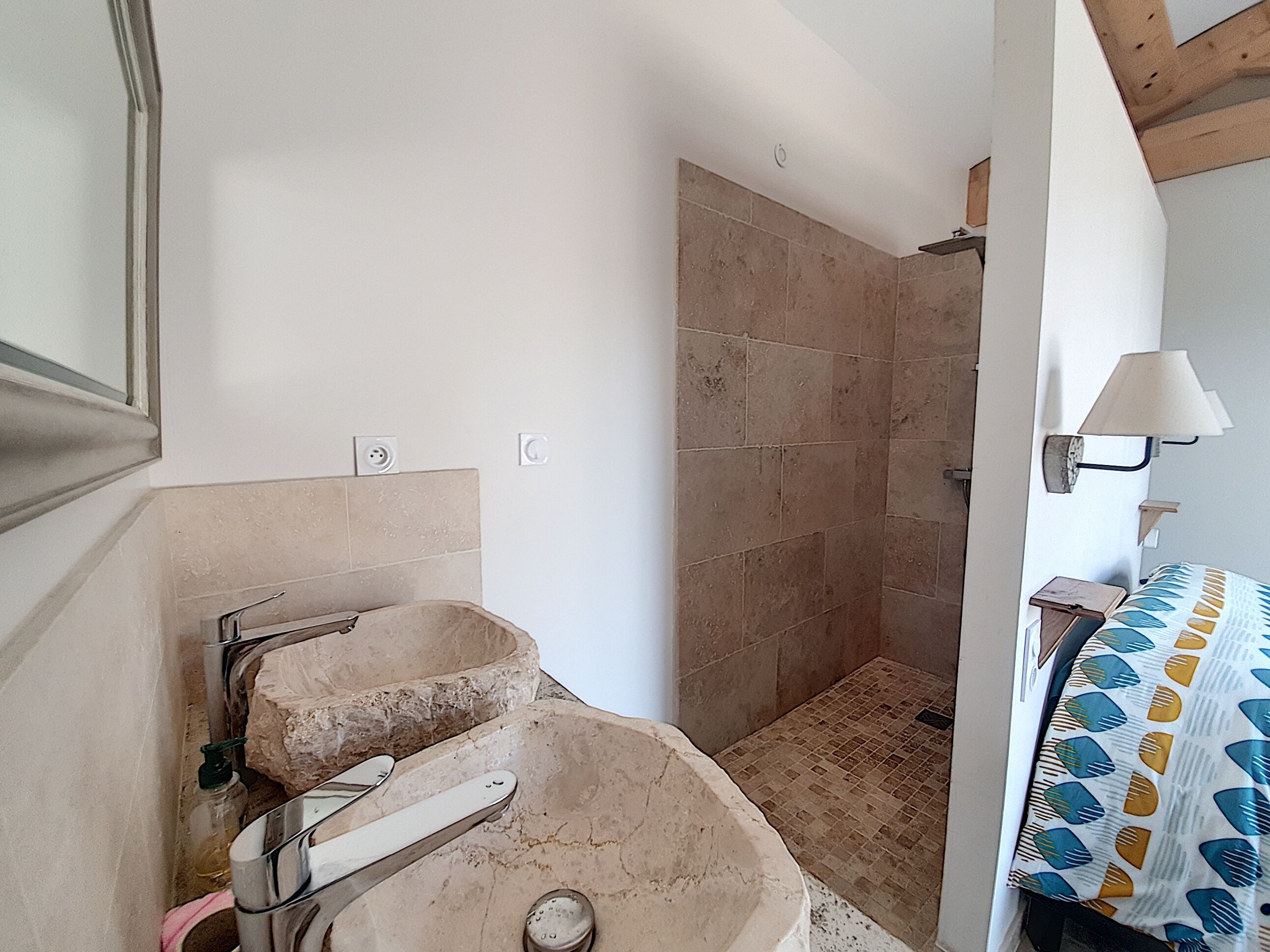 Un baño con lavamanos de piedra, zona de ducha y cama con colcha estampada.