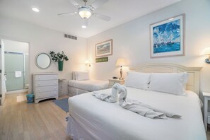 Ein Hotelzimmer mit Bett, Nachttisch, Lampe, Wandgemälde, Deckenventilator und einem Badezimmer mit Dusche und Toilette.