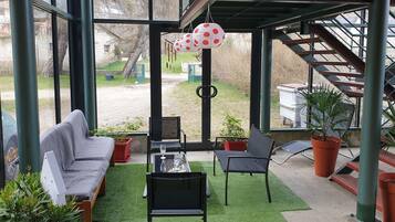 Un espace extérieur aménagé, abrité et doté d’un canapé, de chaises et d’une table, entouré de plantes en pot, avec vue sur un jardin et un bâtiment en arrière-plan.
