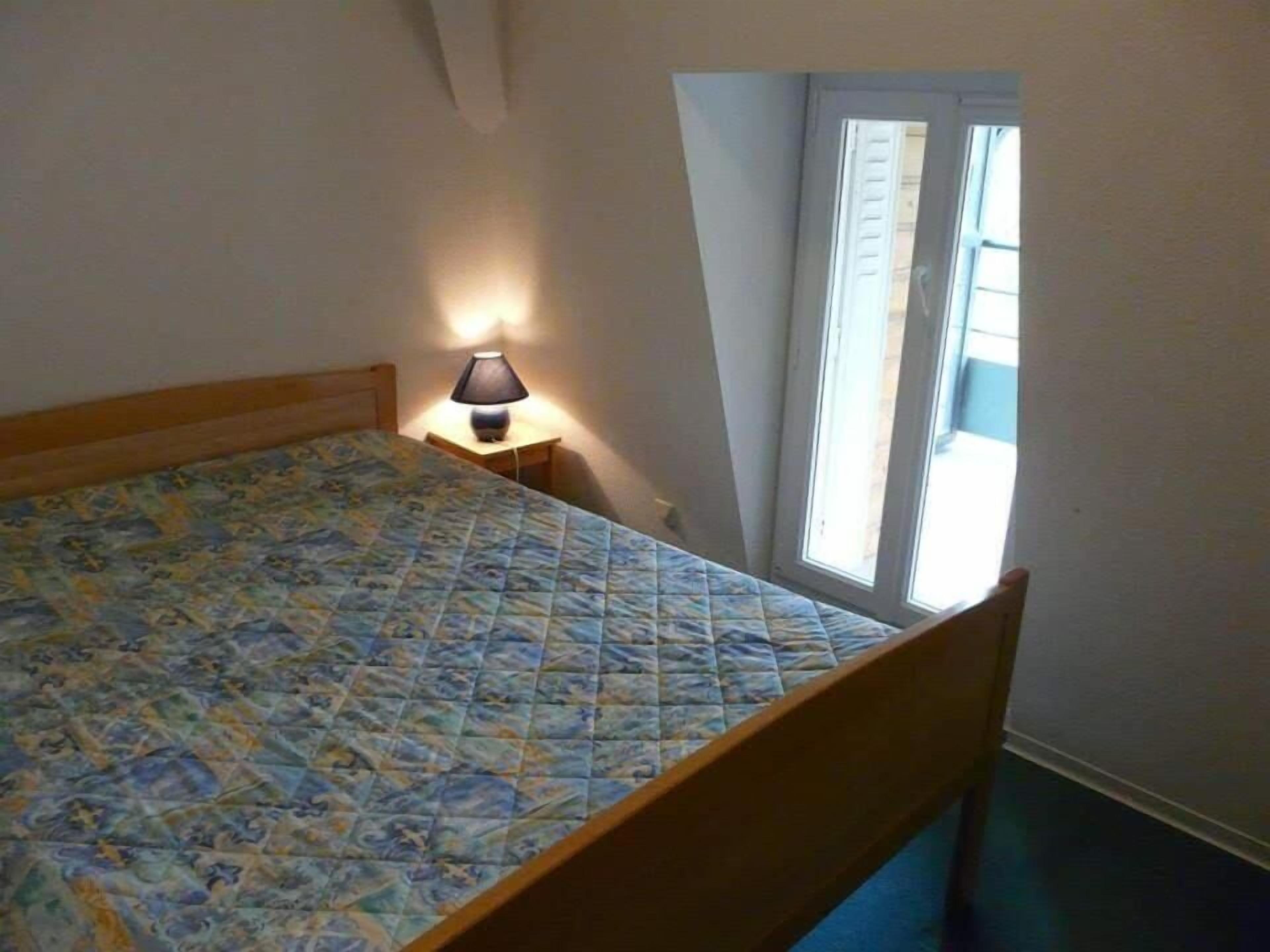 A bedroom with a bed, a nightstand, a lamp, and a window.