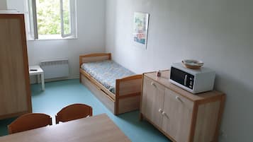 Une petite pièce bien rangée, avec un lit simple, un bureau, une chaise et un four à micro-ondes posé sur un meuble.