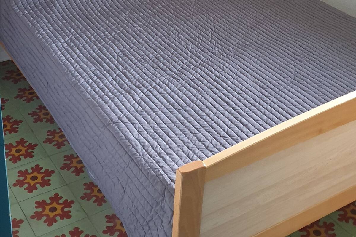 A single bed with a wooden frame and a grey quilted mattress, a framed picture on the wall, and a patterned floor.