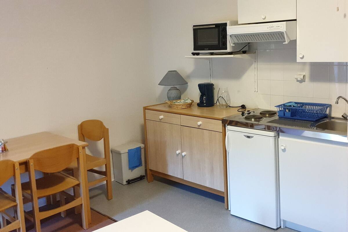 A compact kitchen with a microwave, a sink, a small refrigerator, a wooden table with chairs, and a blue basket.