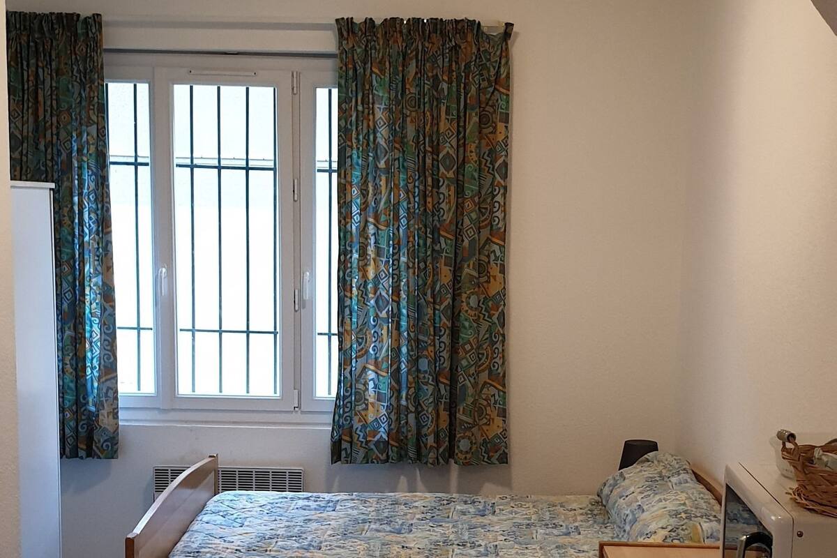 A small, single-room apartment with a bed, a wooden dresser, a microwave, and a window with curtains.