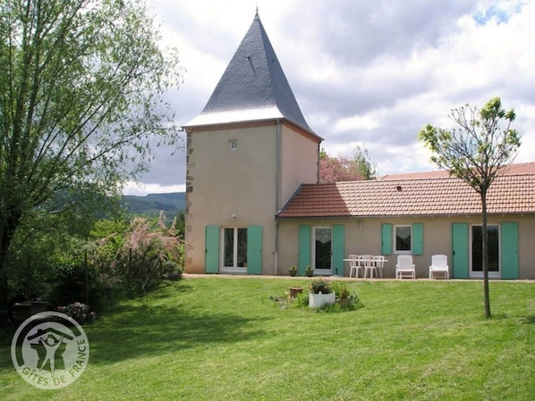 A two-story building with a conical roof, a flat-roofed section, and a small tower. There is a grassy lawn in front with a few potted plants and outdoor furniture.