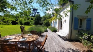 A wooden deck with a round table and chairs, surrounded by lush greenery and a white building with blue shutters.