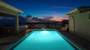 Piscina com vista para o pôr do sol sobre o oceano, rodeada por espreguiçadeiras e uma área coberta.