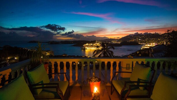 Terraço com cadeiras e uma mesa, com vista para o horizonte costeiro ao entardecer, com edifícios iluminados.