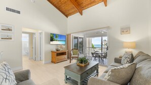 A spacious living room with a wooden ceiling, a television, a coffee table with a plant, and a view of the ocean through a sliding glass door.