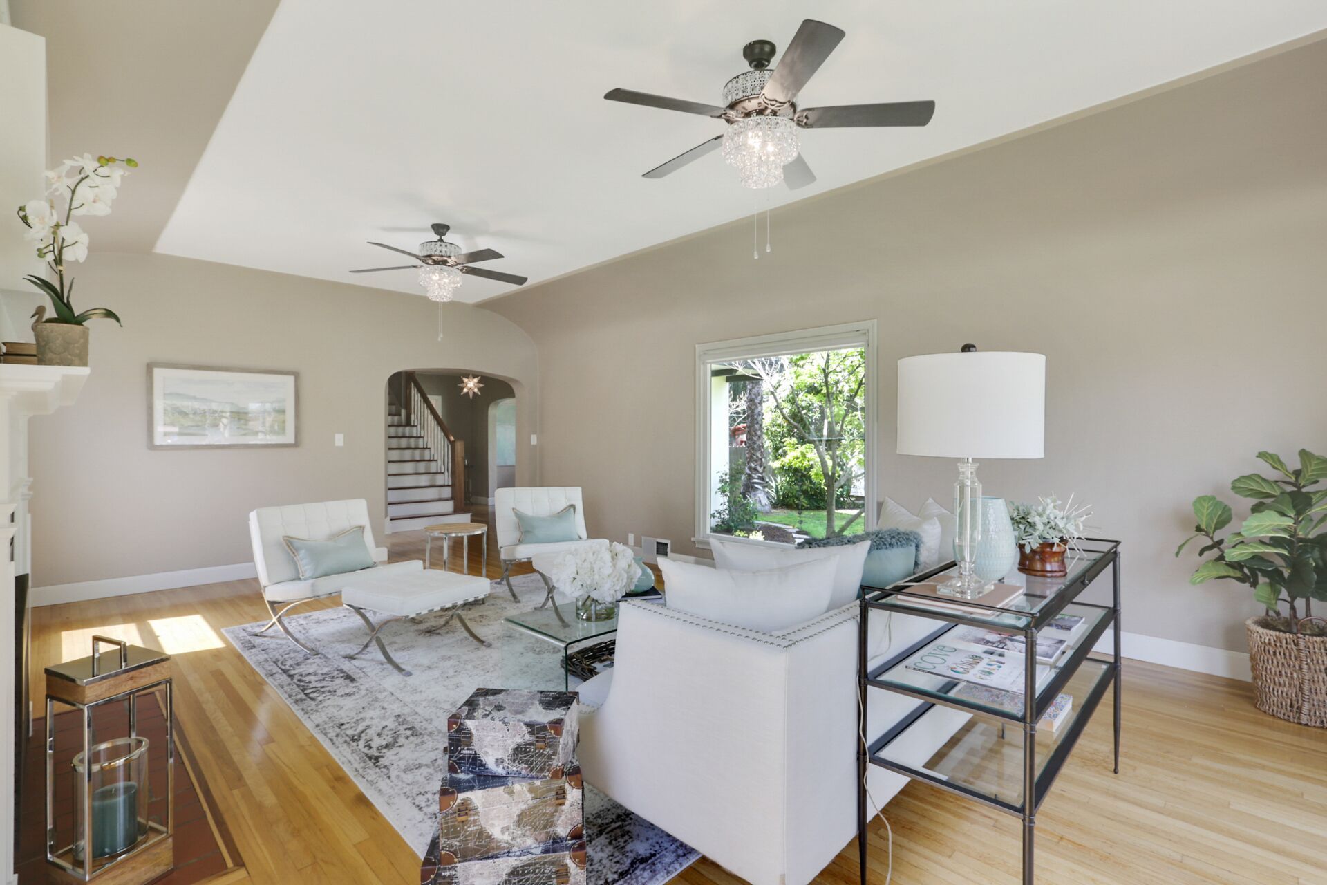 A spacious living room with a high ceiling, two ceiling fans, a large window with a view of greenery, a white sofa set, a glass coffee table, and a side table with a lamp.