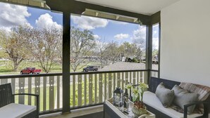Un balcon donnant sur un quartier résidentiel, une chaise en osier, un canapé et un plateau avec une bouteille et des fleurs.